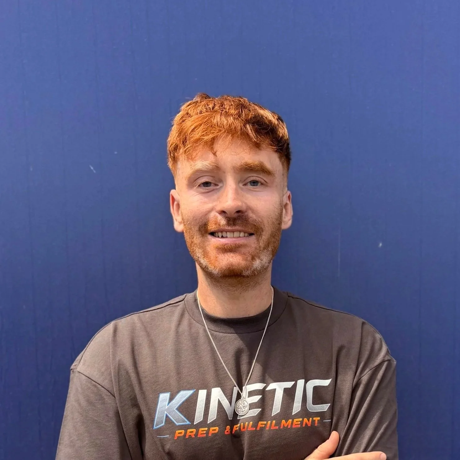 A young man with red hair, beard, and blue eyes smiling, standing against a solid blue background, wearing a gray t-shirt with "KINETIC" and "PREP & FULFIMENT" printed on it, and a silver necklace.