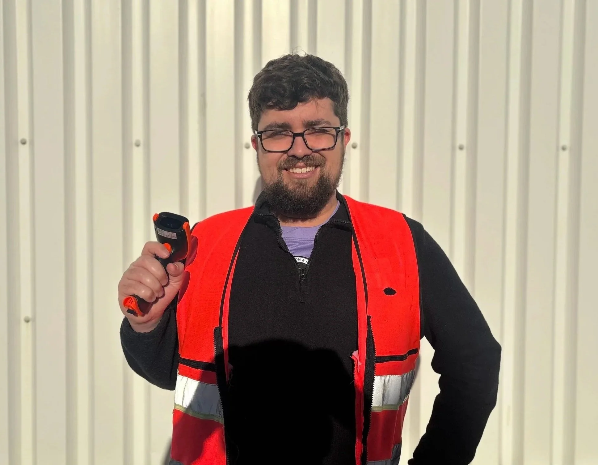 A man with dark, curly hair, glasses, and a beard, looking to his left, wearing a black sweater and standing in a room with a plant in the background.