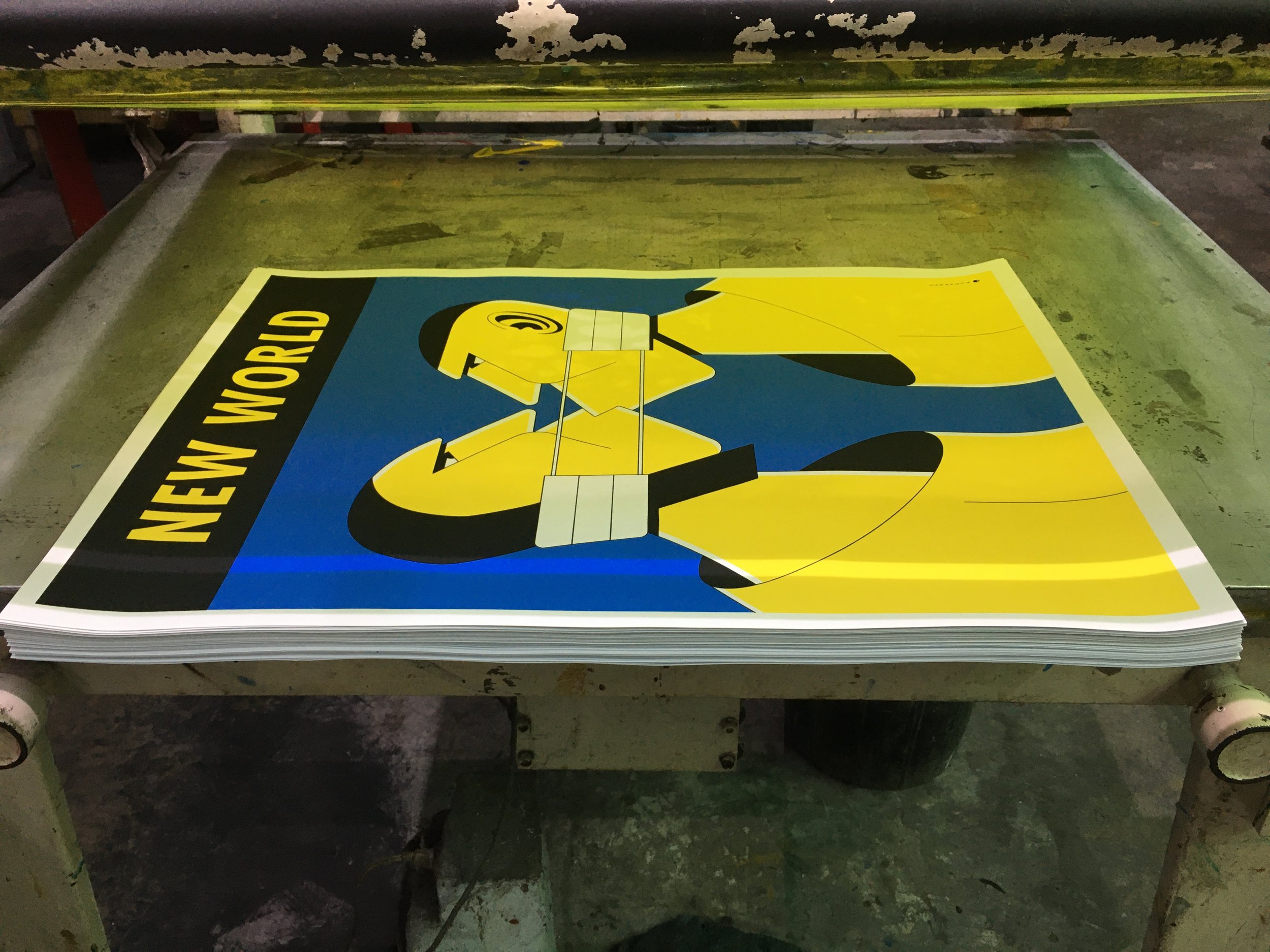 A stack of posters on a work table, featuring a drawing of a person in yellow and black with the text 'NEW WORLD' in bold black and yellow letters.