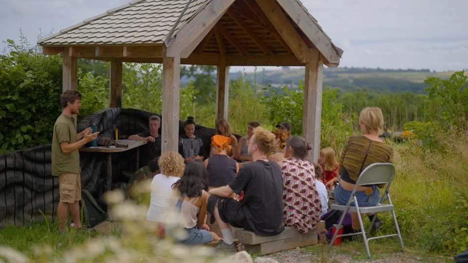 A group of people gathered outdoors under a small wooden gazebo, listening to a young man speaking. The group includes adults and children sitting on benches and a folding chair, with a natural landscape of trees and fields in the background.