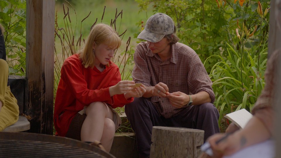 A young girl and an older man sitting outdoors on a wooden bench, both looking at a small object the girl is holding.