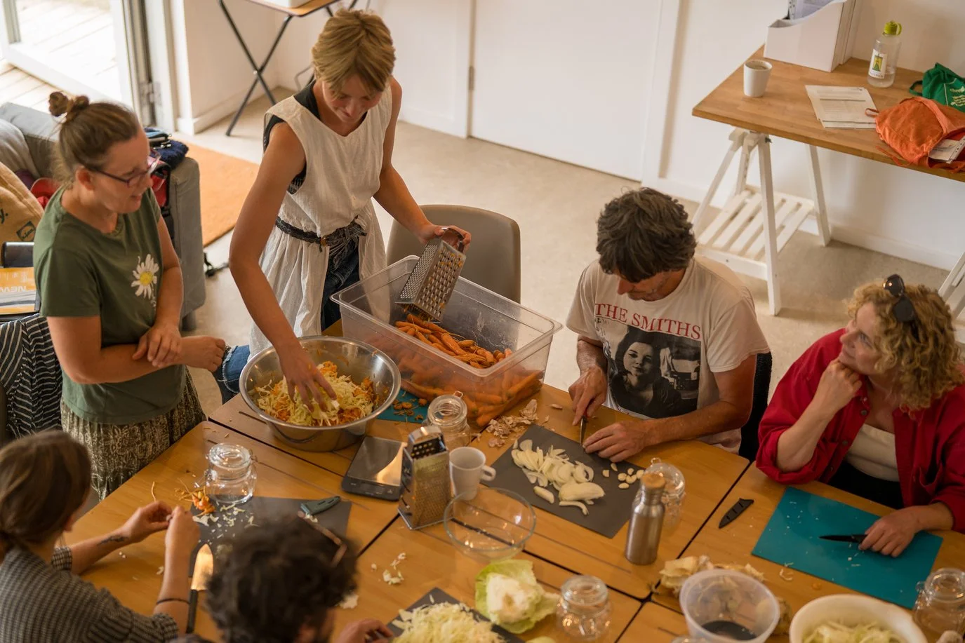 People preparing food in a kitchen, chopping vegetables and mixing ingredients around a table with various food items and kitchen tools.