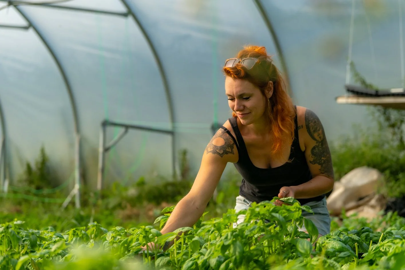 A woman with red hair and tattoos on her arms, wearing a black tank top, tending to green plants inside a greenhouse.