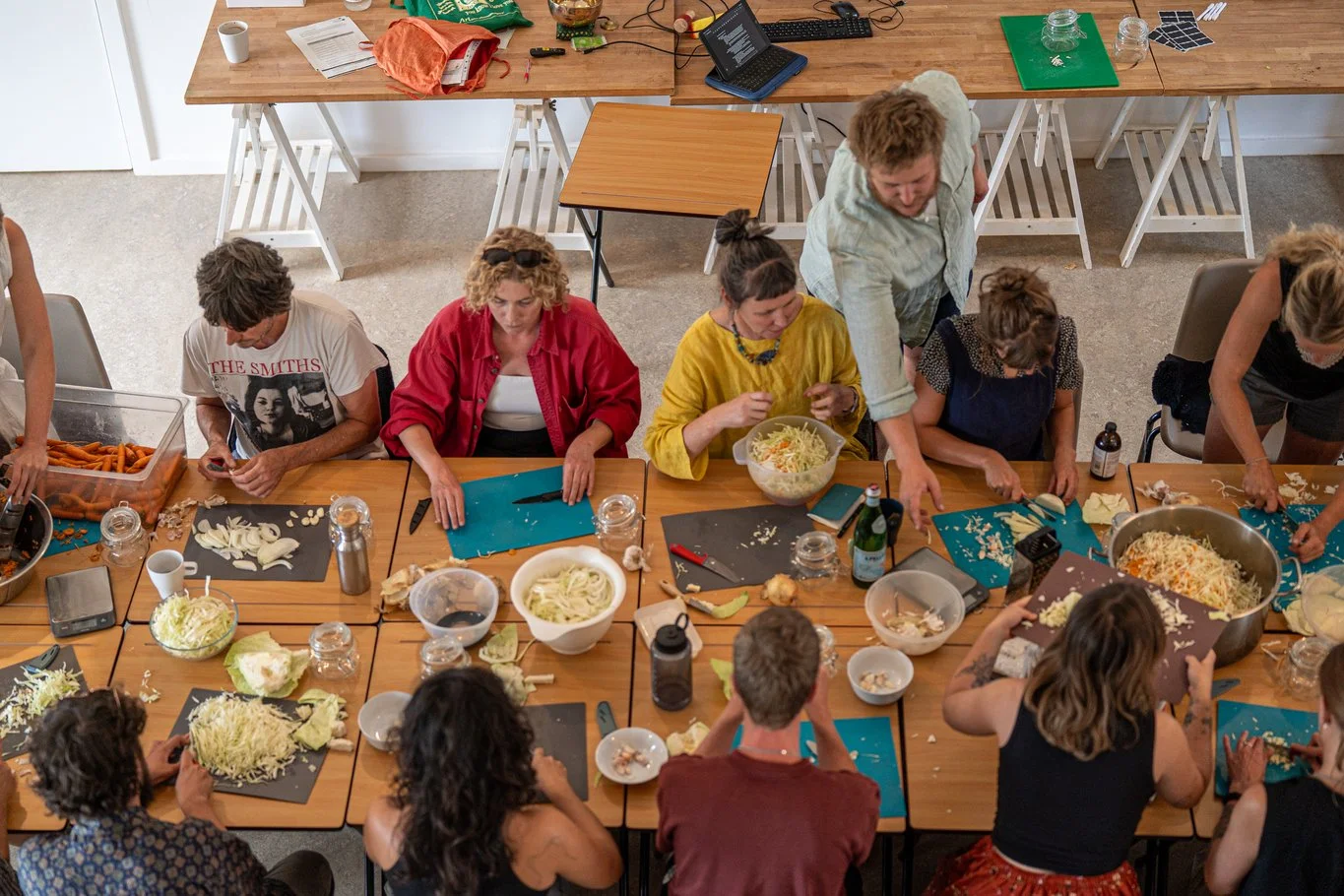 Group of people preparing food together at a long wooden table, chopping vegetables, with bowls of shredded cabbage and other ingredients, surrounded by kitchen utensils and glasses.