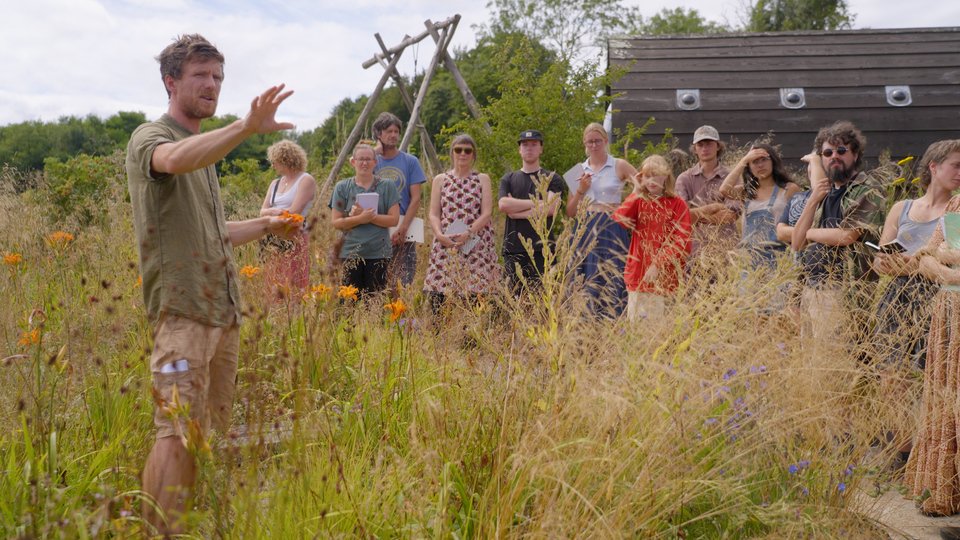 A man is speaking to a group of people sitting outdoors in a grassy field during daytime.