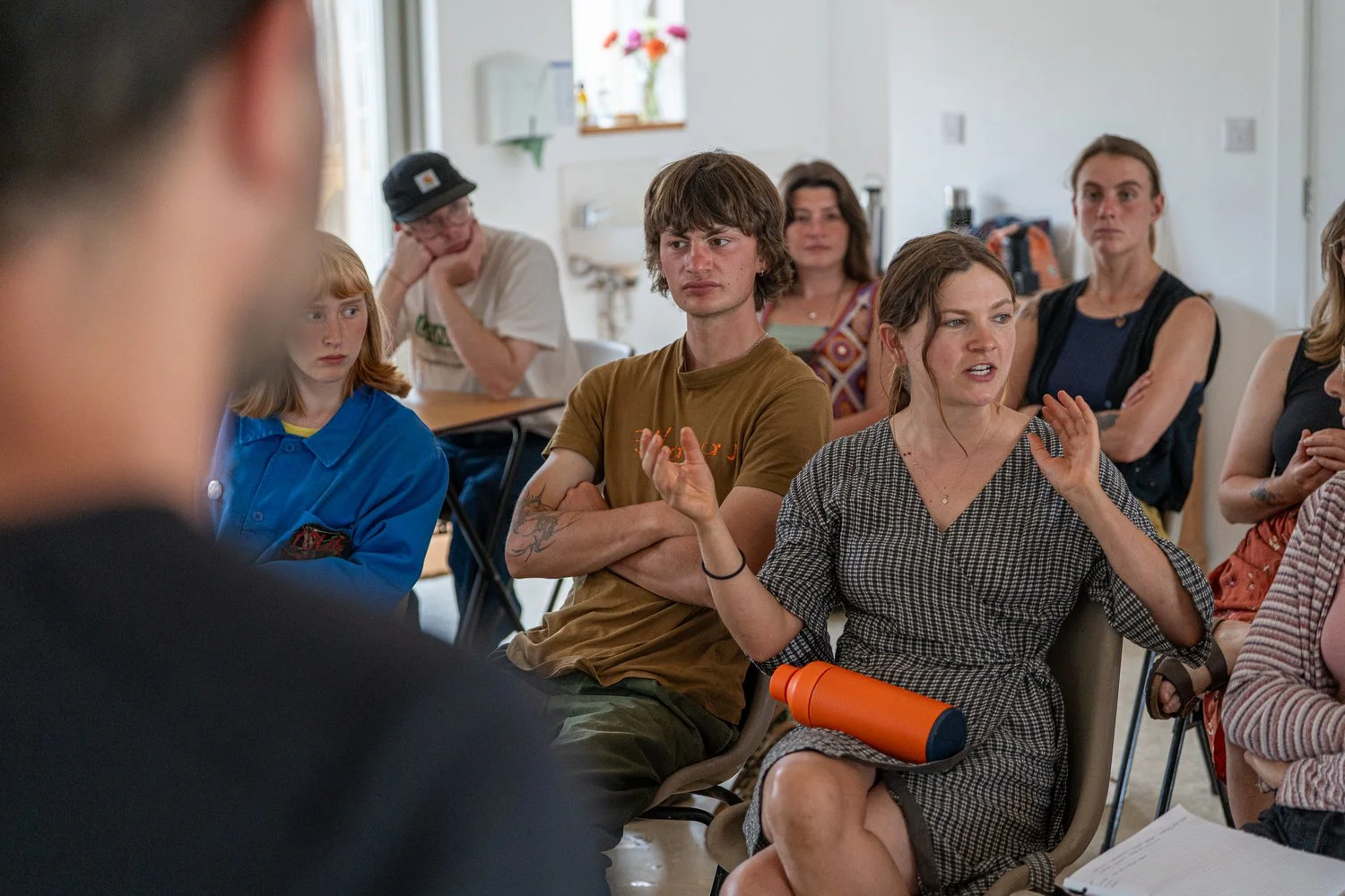 People attending a discussion or meeting in a small room, with a woman speaking and gesturing while others listen.