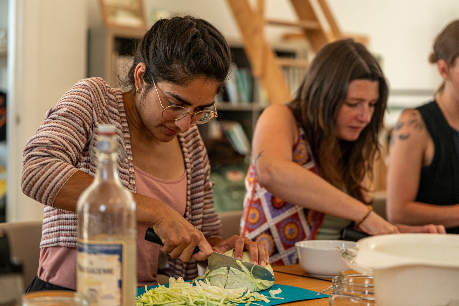 Three women chopping cabbage together in a kitchen with wooden shelves and books in the background.