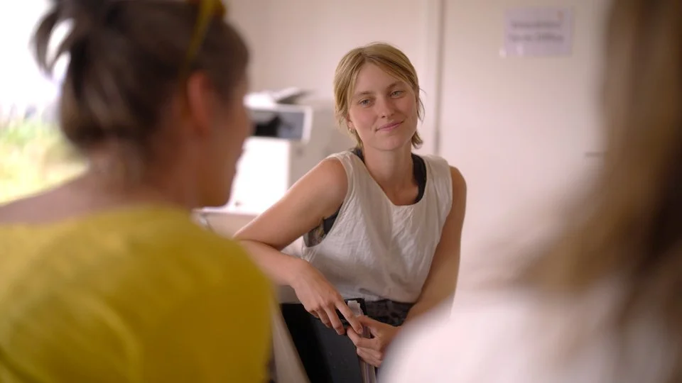 A young woman with short blonde hair sitting in a room with a slight smile, wearing a white sleeveless top, and engaging in conversation with two other women, one in a yellow top and the other blurred in the foreground.