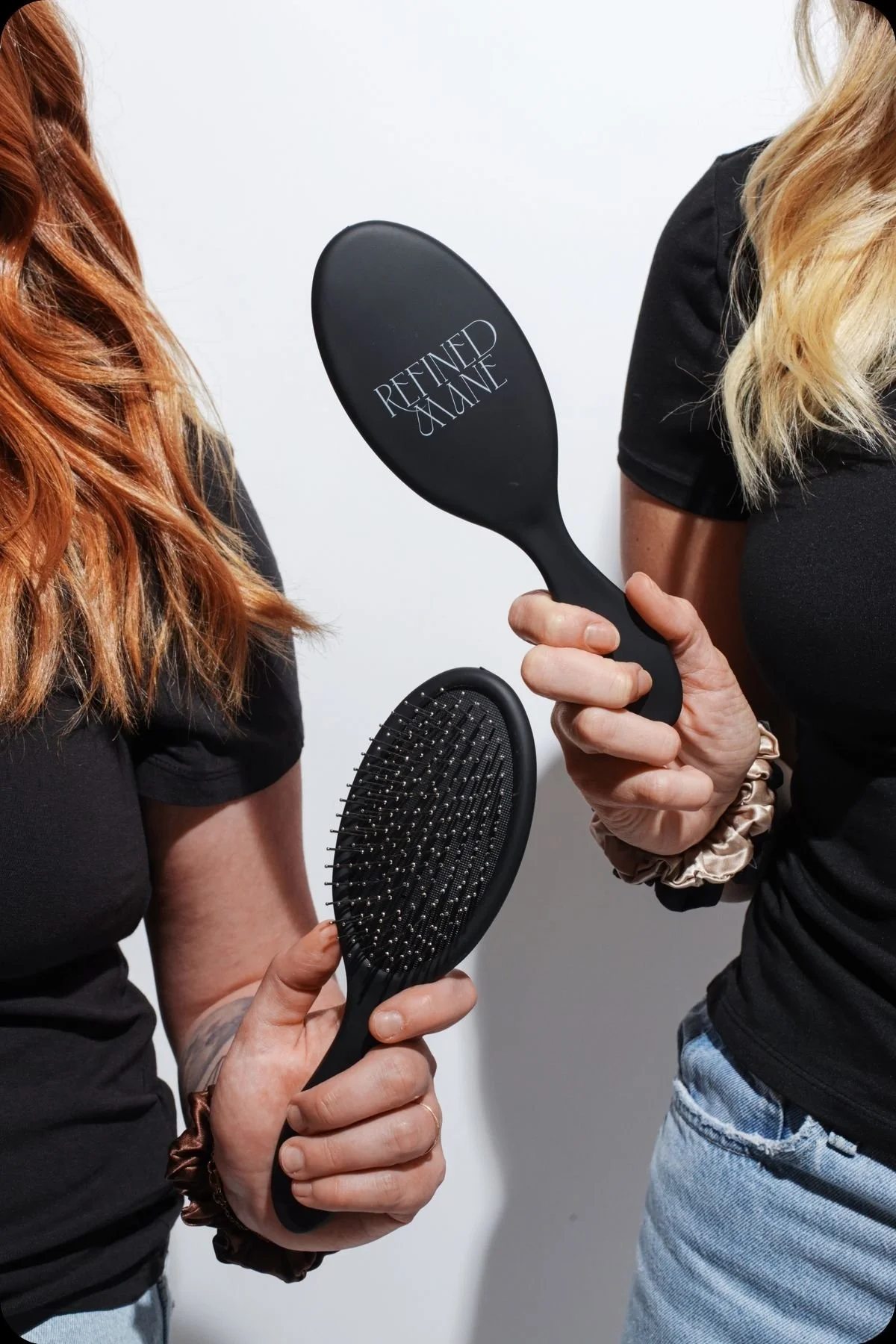 Close-up of two Refined Mane hair brushes showing front and bristles beside blonde and auburn hair.
