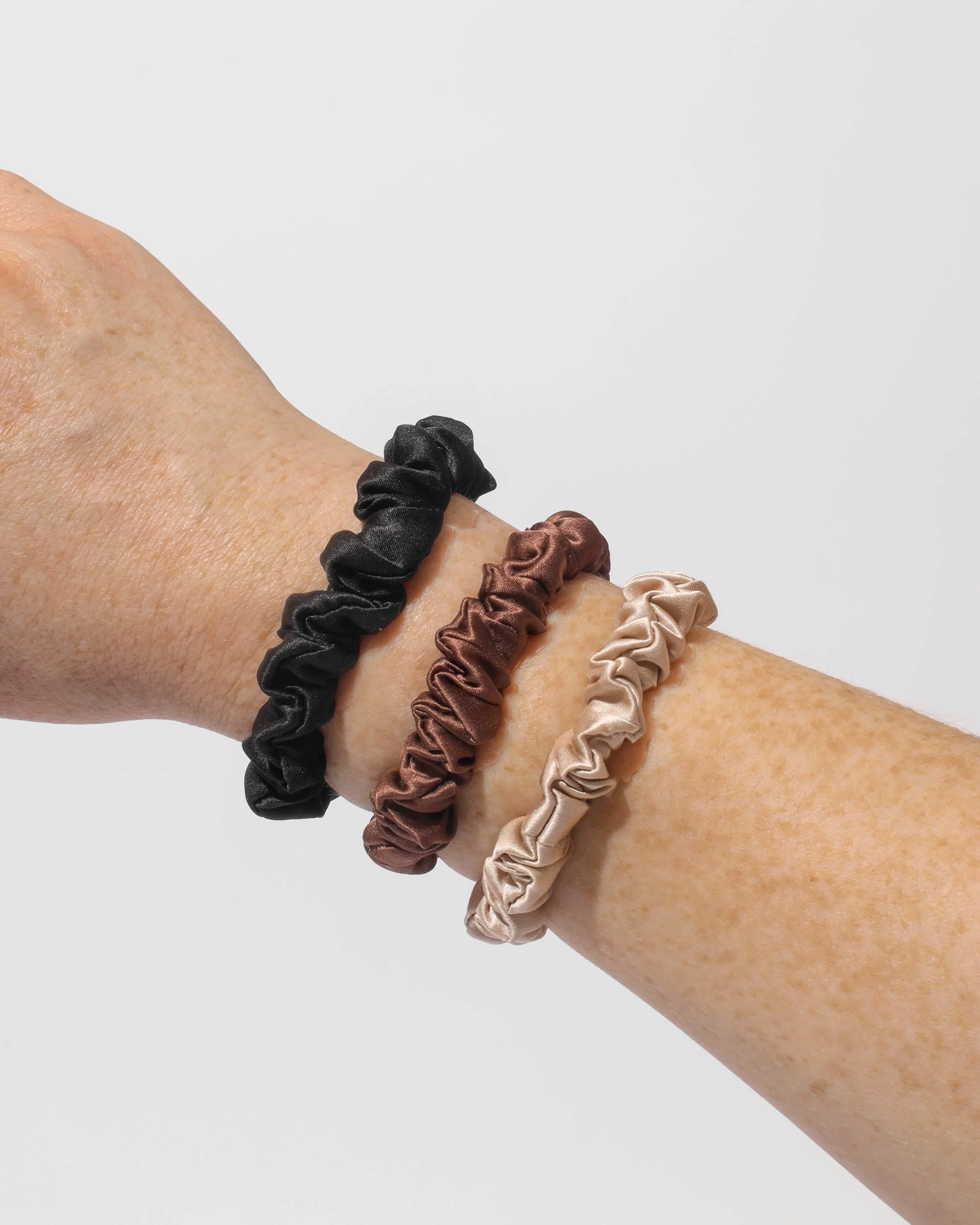 3 small Refined Mane silk scrunchies in champagne, chocolate, and onyx worn on wrist.