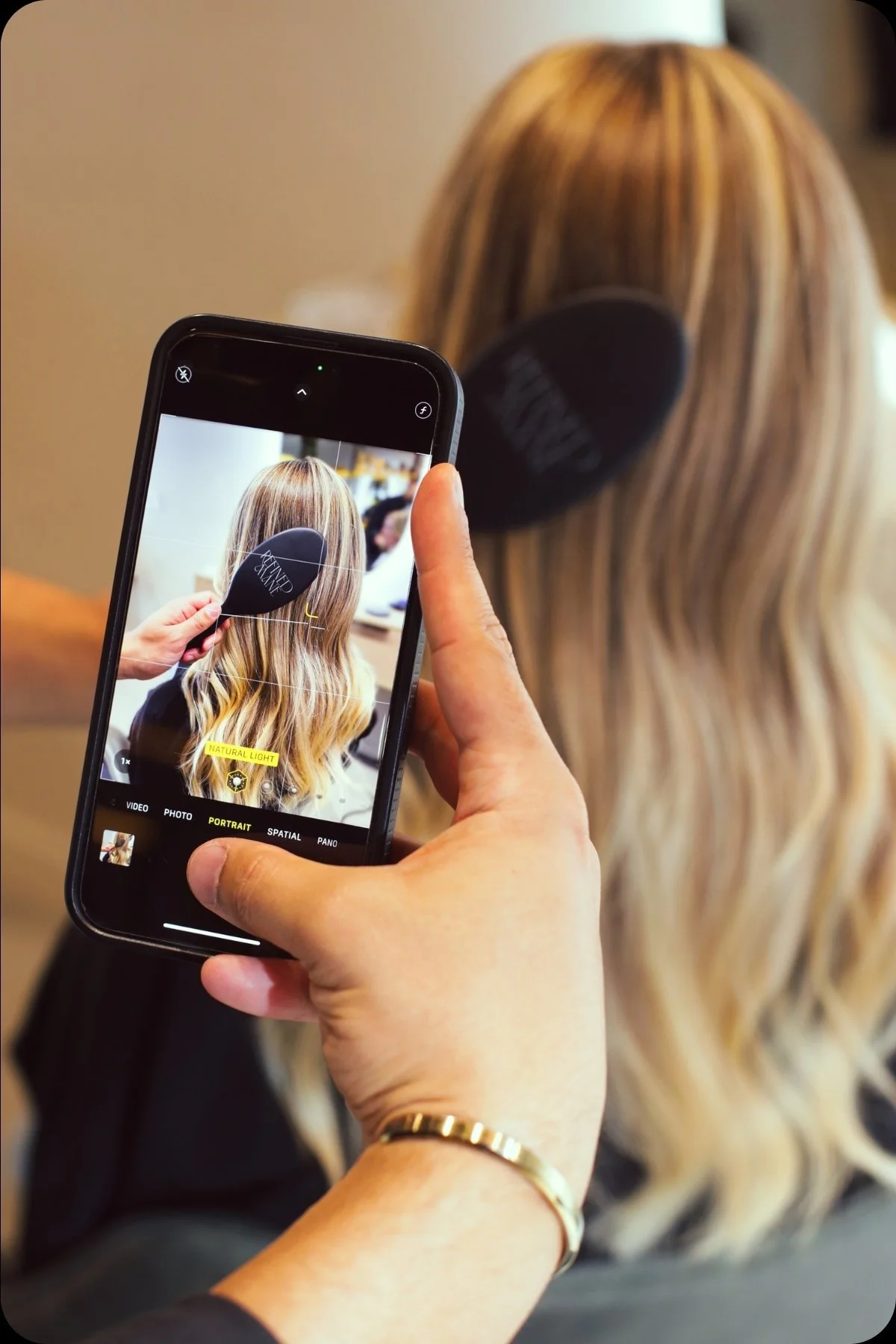 Stylist photographing blonde hair styled with a Refined Mane brush in a salon setting.