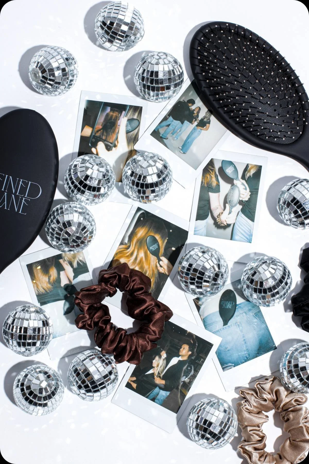Disco balls, polaroid photos, Refined Mane silk scrunchies, and hair brushes arranged on a white surface.