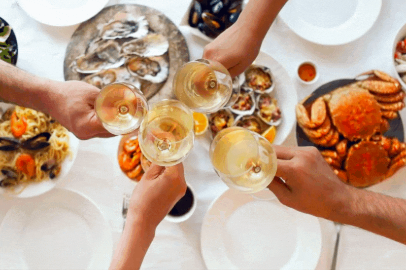 Four people clinking glasses of white wine over a table filled with seafood dishes including crab, oysters, and mussels.