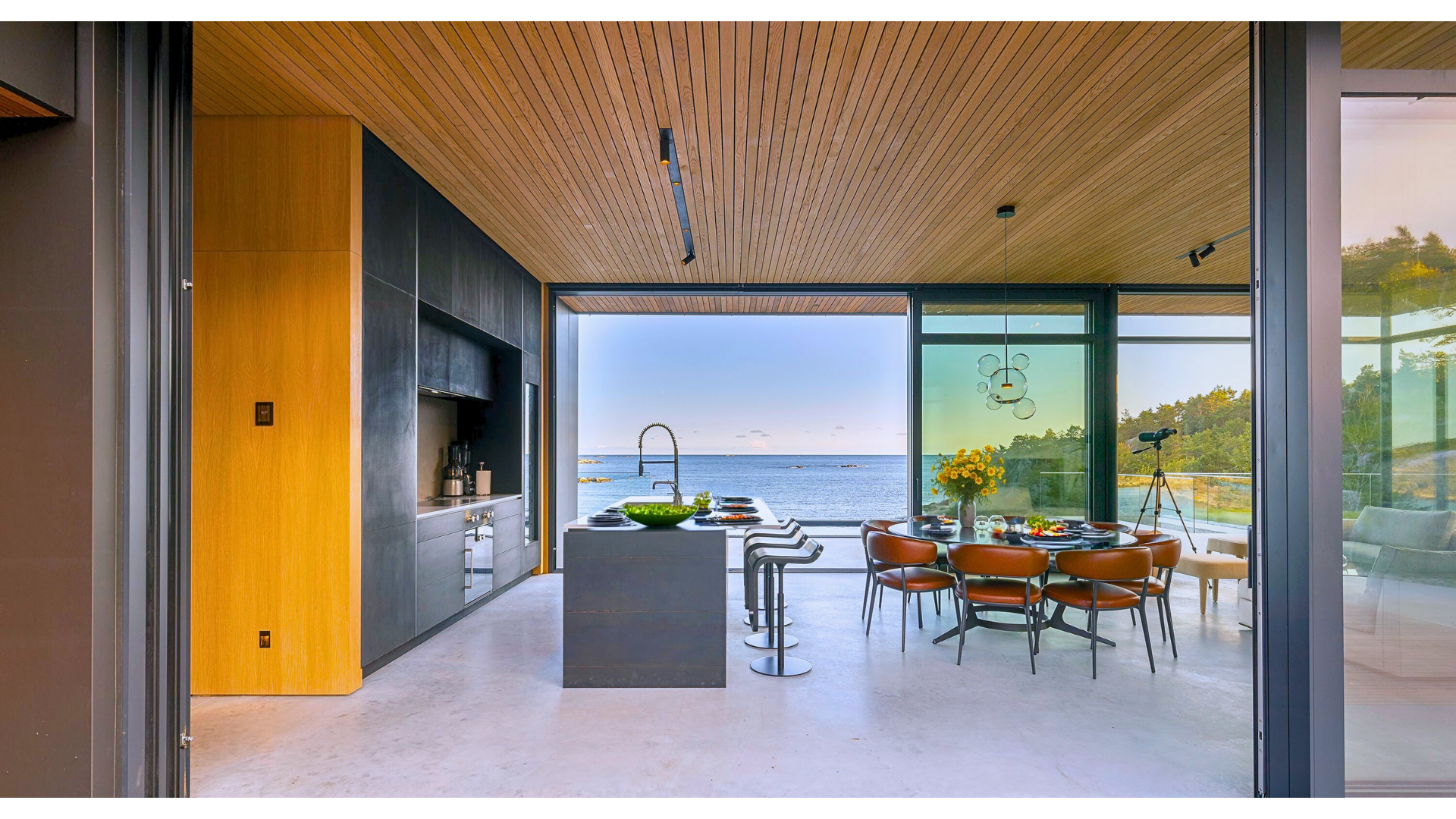 Modern open-concept kitchen and dining area with large glass windows overlooking the ocean, wooden ceiling, black cabinetry, round dining table with orange chairs, island with bar stools, and telescope on a balcony.