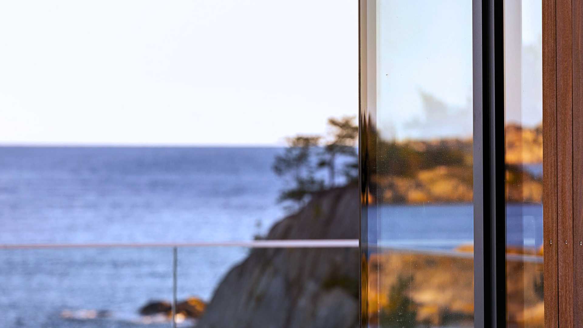 Close-up of a glass window reflecting a rocky coastline and trees during sunset, with part of a wooden wall visible.