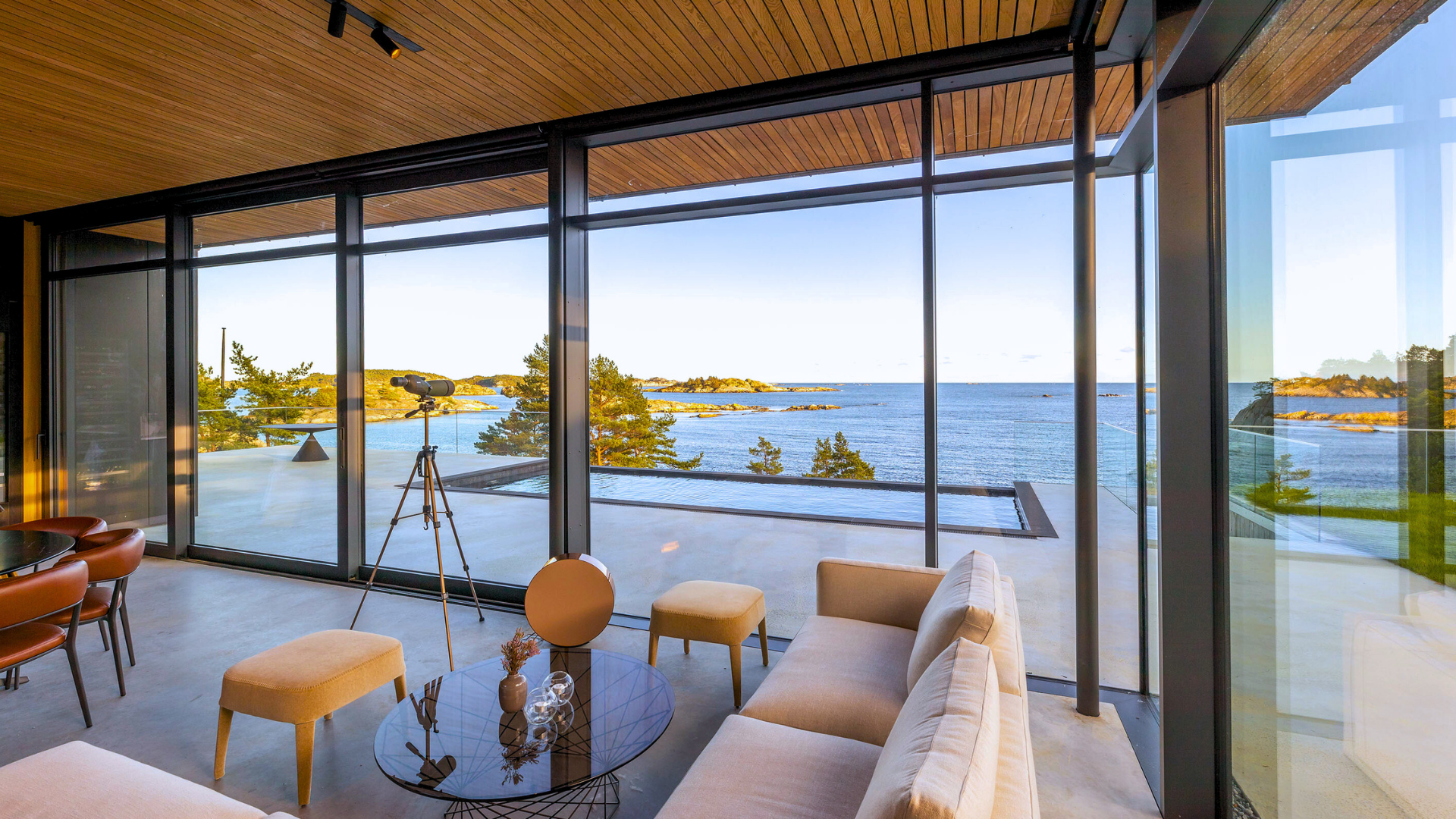 Living room with floor-to-ceiling windows overlooking the ocean, with a telescope on a tripod, beige sofa, yellow stools, and a round glass coffee table with decorative items.