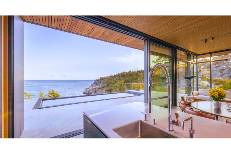 Modern kitchen with ocean view, large open sliding glass doors showing outdoor patio and rocky shoreline, interior includes a round dining table with a potted sunflower, and a sitting area with a telescope.