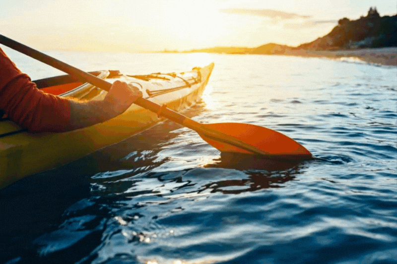 A person kayaking on calm water during sunset with a distant shoreline and hills in the background.