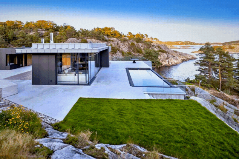 Modern house on a rocky hillside overlooking a river with surrounding trees and landscape, featuring large glass windows and a minimalist design.
