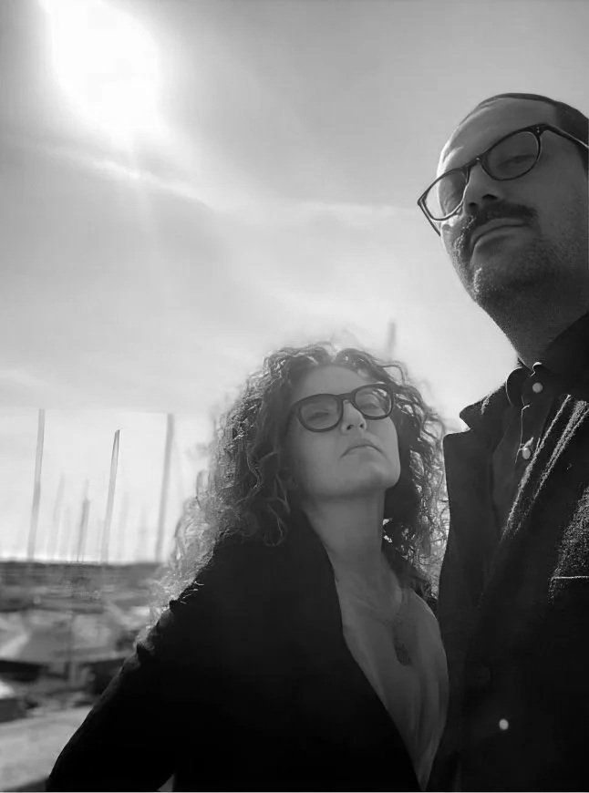 Due persone, un uomo e una donna, indossano occhiali e si scattano una foto all'aperto con il cielo e alcune barche sullo sfondo.