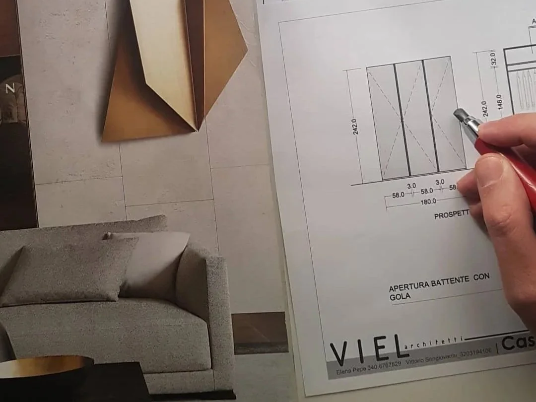 Pianta di un progetto architettonico con dettagli, dimensioni e sezioni, con una mano che tiene una penna. In basso a sinistra, un divano grigio e una ciotola dorata sul tavolo.