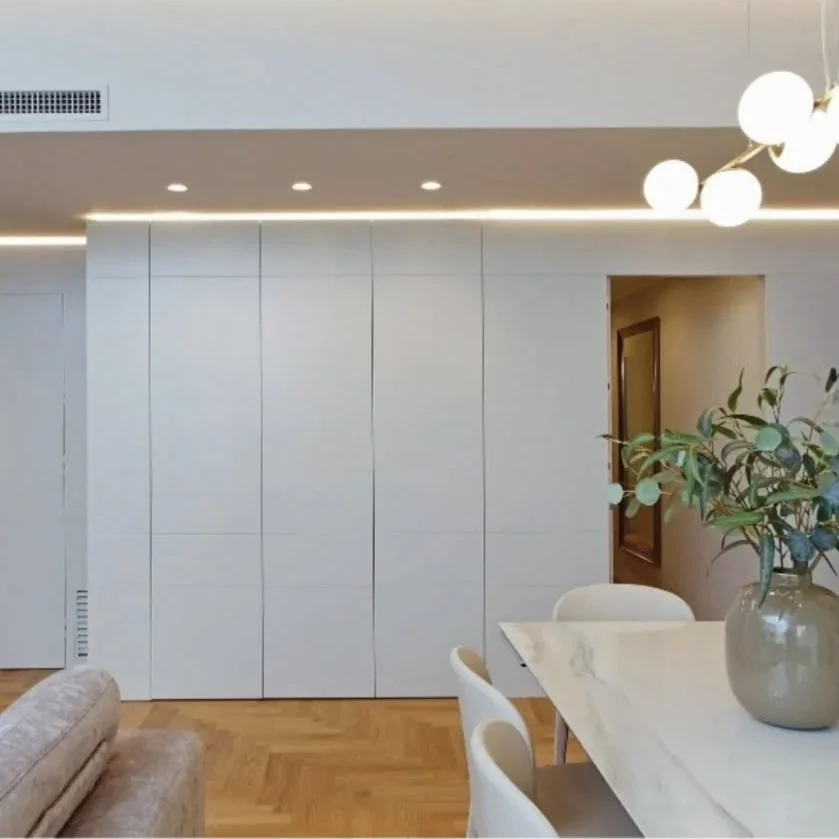 Interno moderno con parete bianca, tavolo con vassoio e sedie, lampadario e pianta decorativa.