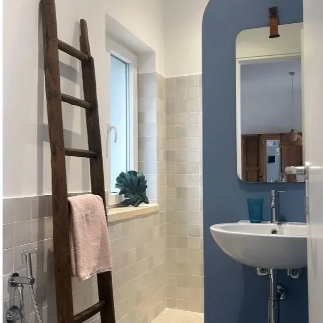 Bagno con pareti rivestite in piastrelle beige, una finestra, uno specchio sul muro blu, lavabo con un bicchiere blu, vaso con pianta sul davanzale, scala in legno appesa alla parete.