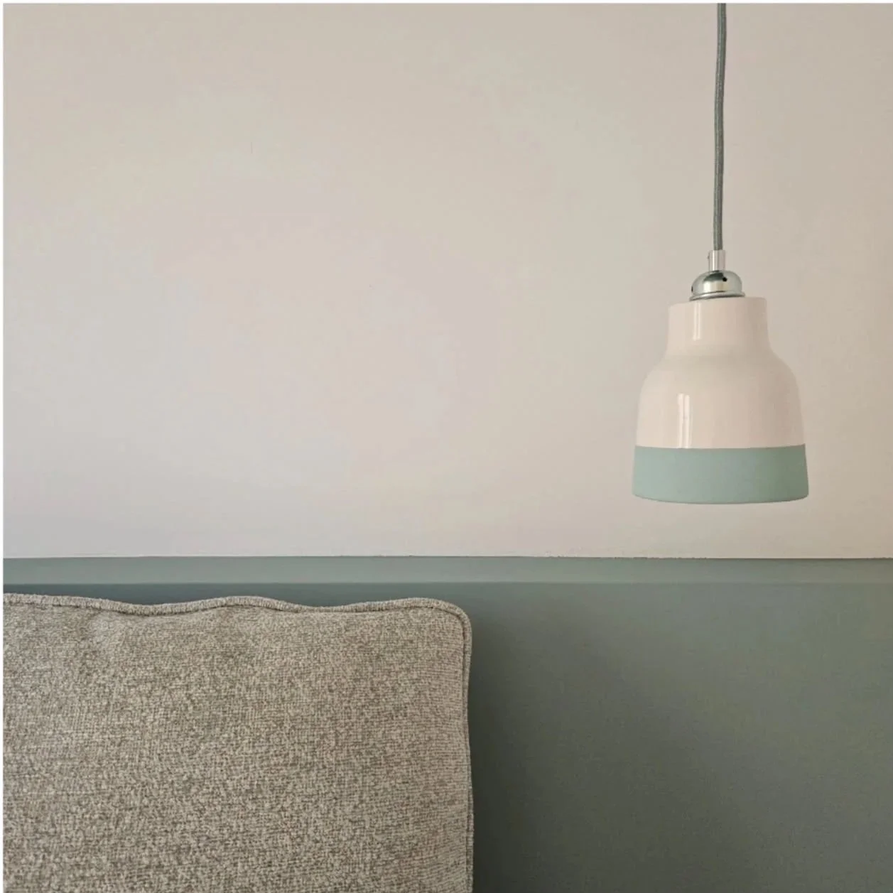 Immagine di una lampada sospesa davanti a una parete di colore chiaro, con un angolo di un divano con rivestimento tessile beige.