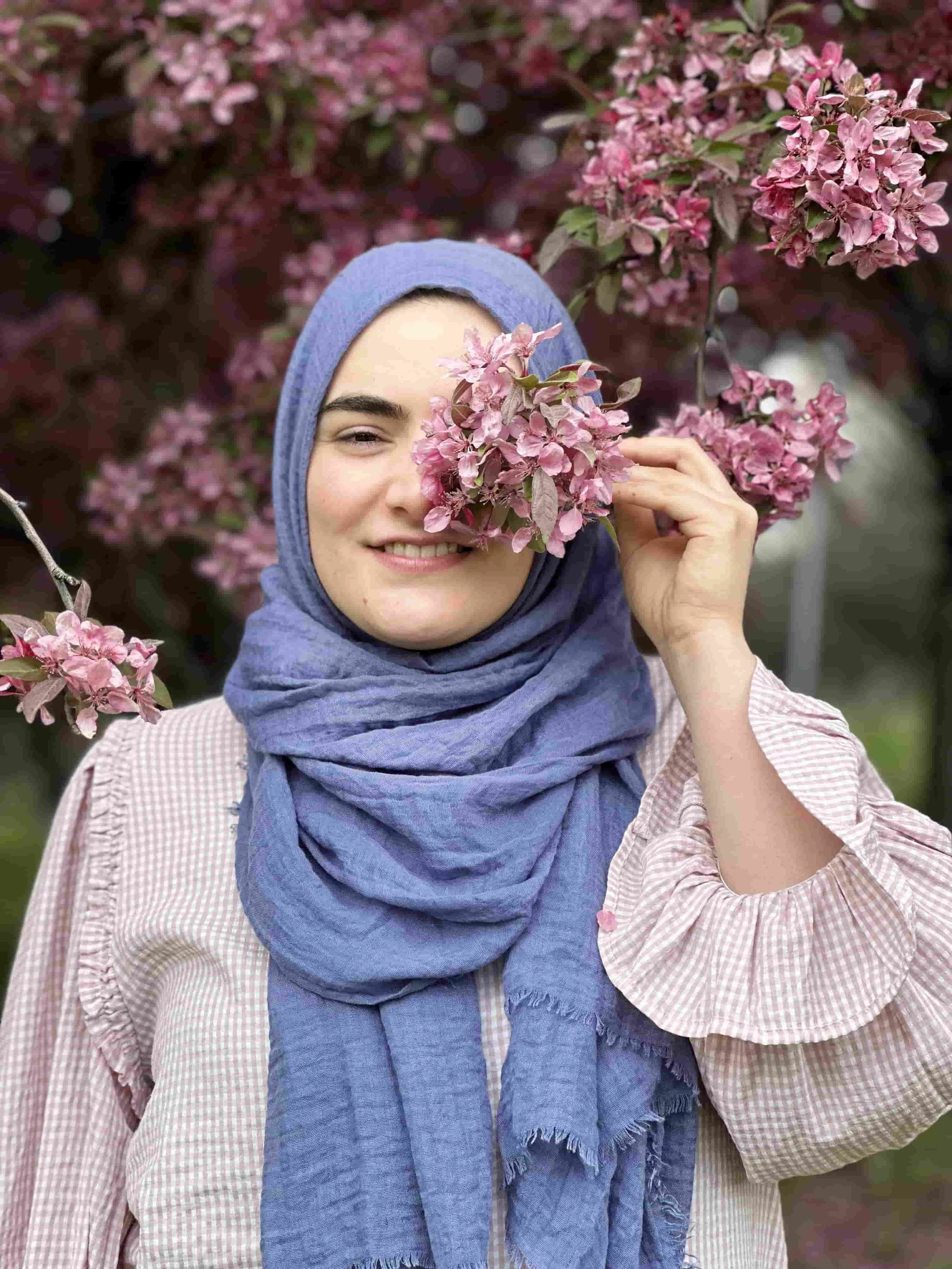 A woman wearing a light blue headscarf and a light pink ruffled blouse stands in front of a tree full of pink blossoms, gently holding a cluster of flowers in front of one eye while smiling | Germanised Gülnur