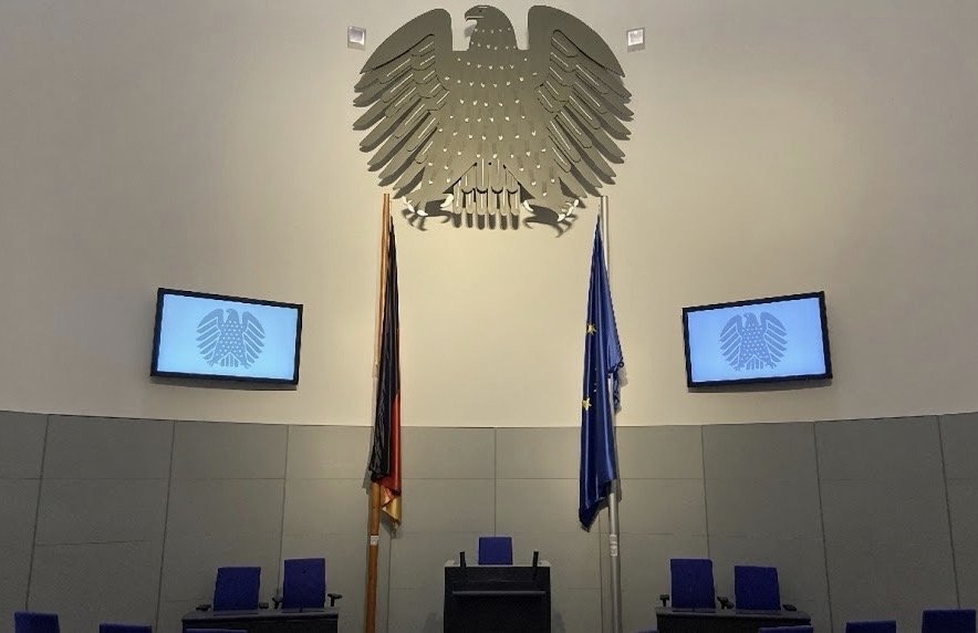 Interior of a German parliamentary chamber with a large silver federal eagle emblem on the wall, German and EU flags beside a central speaker’s podium, blue seats, and two wall screens showing the eagle symbol.