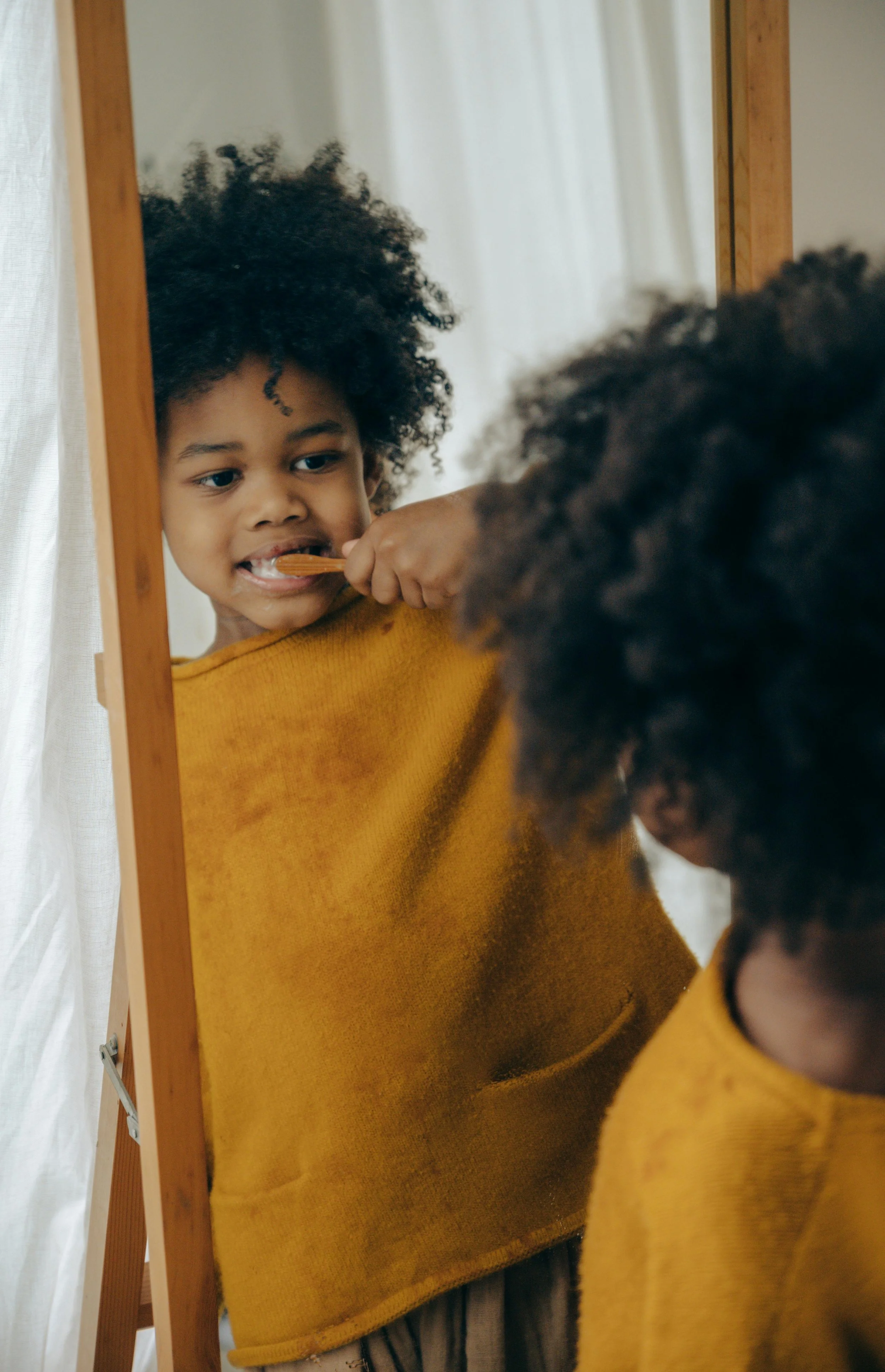 A young girl with curly hair brushing her teeth while looking at her reflection in a mirror.