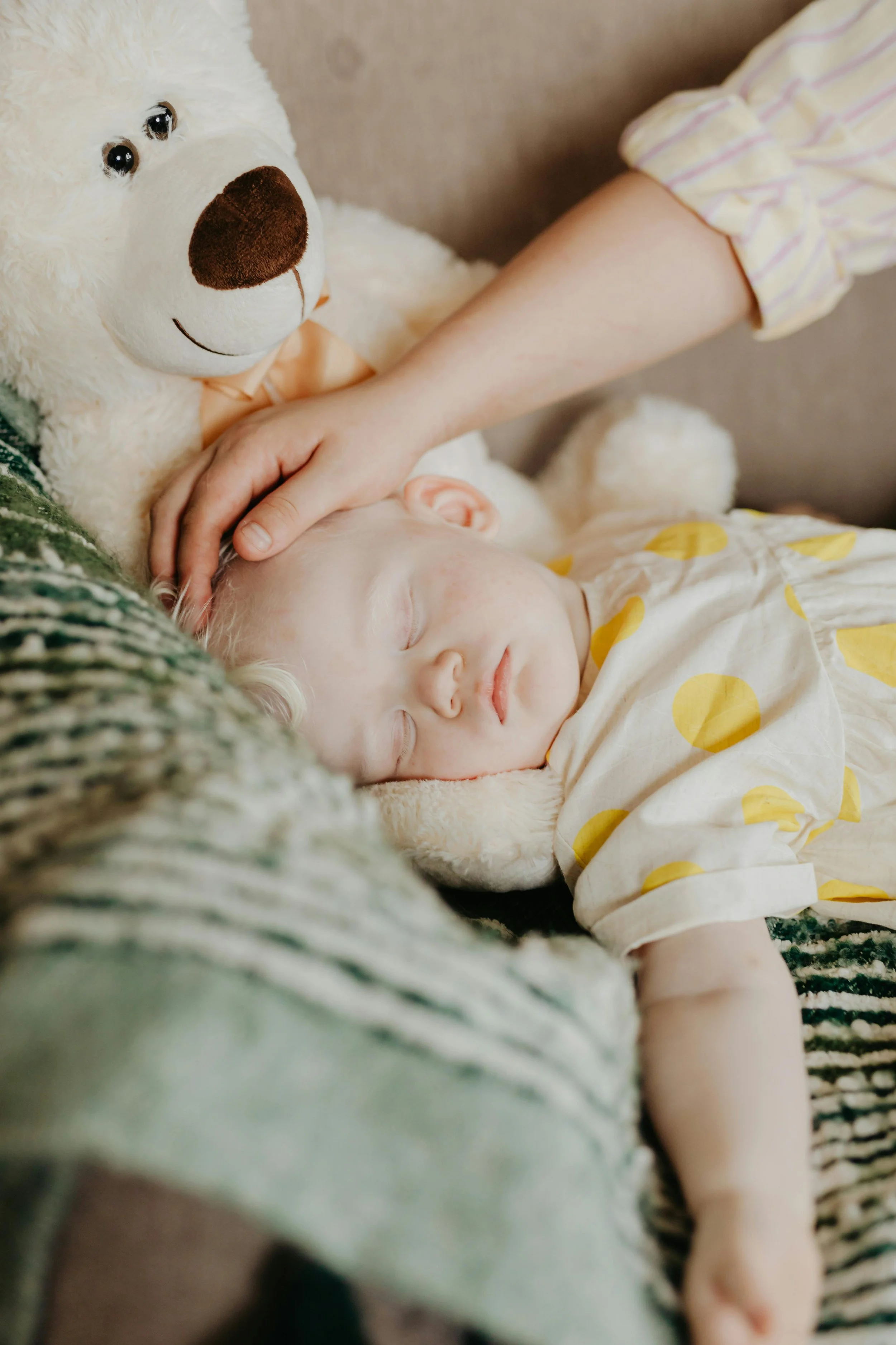 A young child with blonde hair sleeping peacefully on a couch, wearing a cream-colored shirt with yellow polka dots, cuddled with a large white teddy bear with a brown nose, while a person gently pats the child's head.