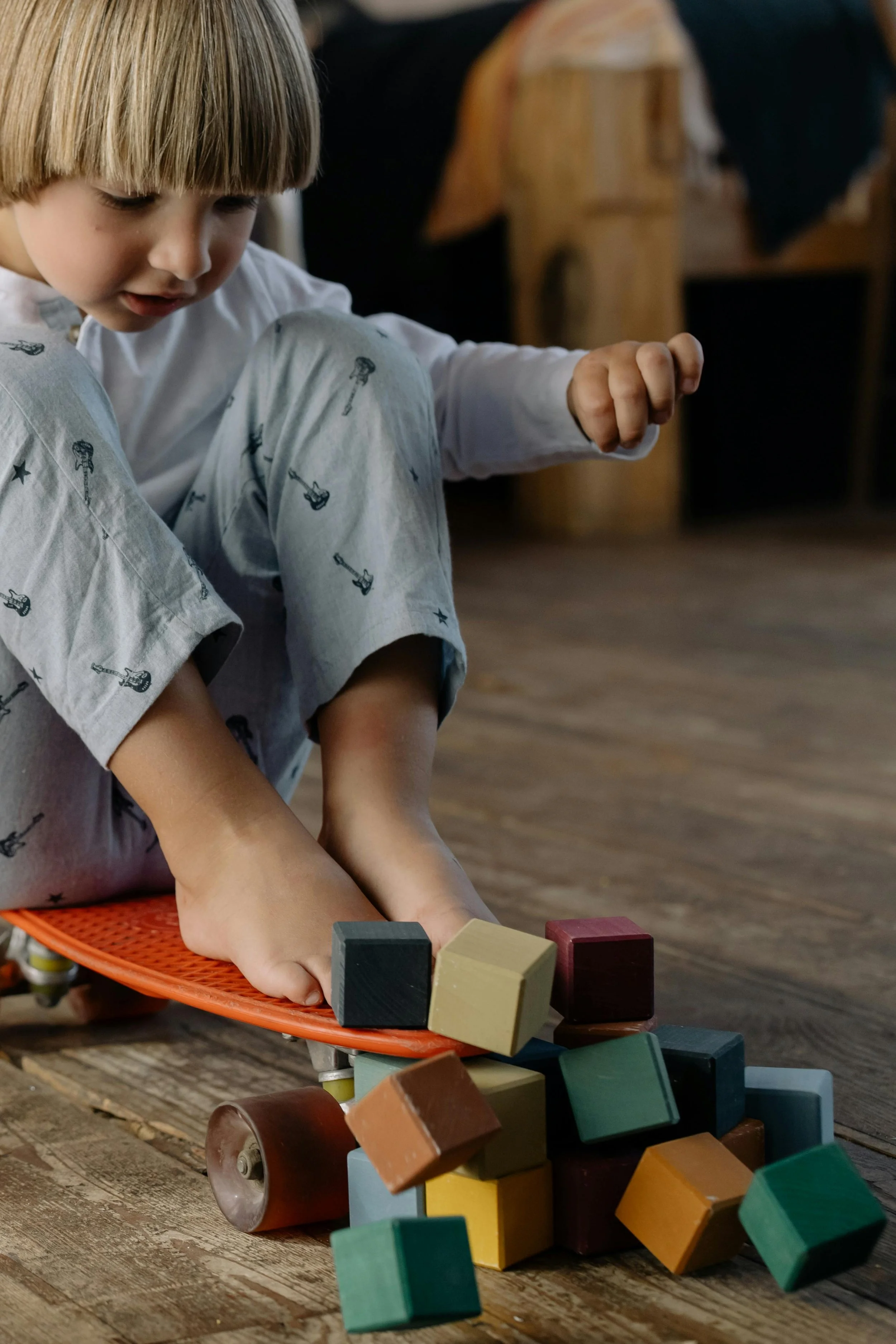A young child in pajamas with a guitar print, sitting on a skateboard, playing with colorful wooden blocks that are spilling on a wooden floor.