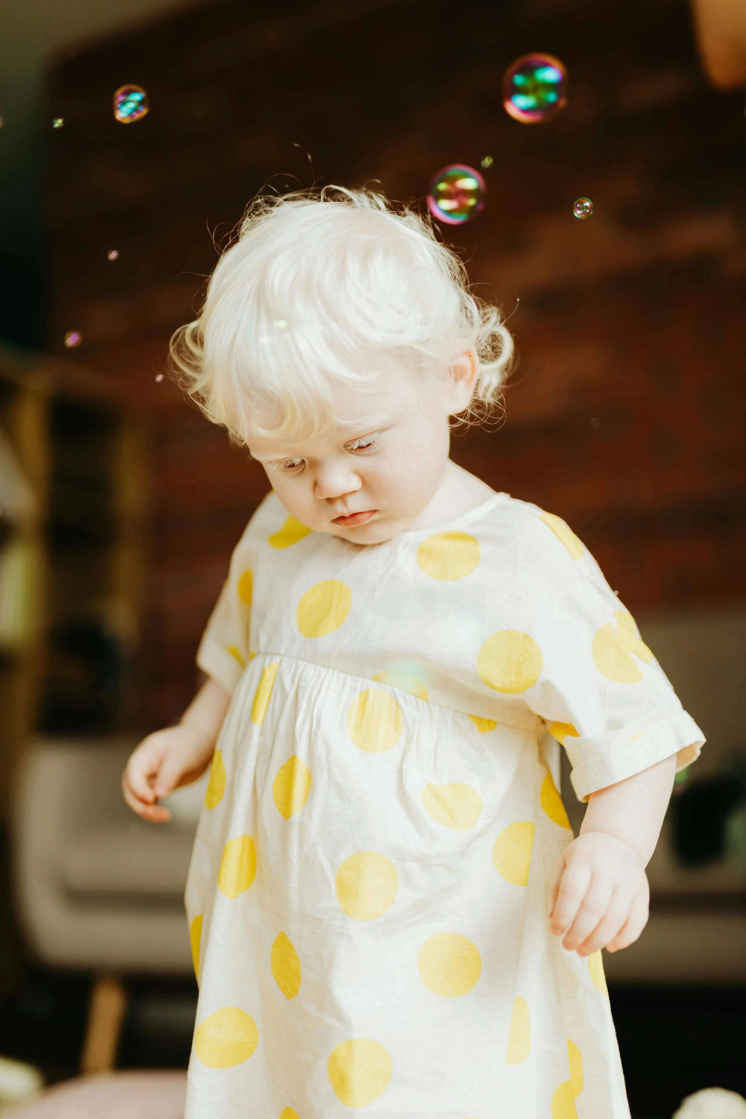 A young child with blonde curly hair wearing a white dress with yellow polka dots, looking down, with colorful bubbles floating around in a cozy indoor setting.