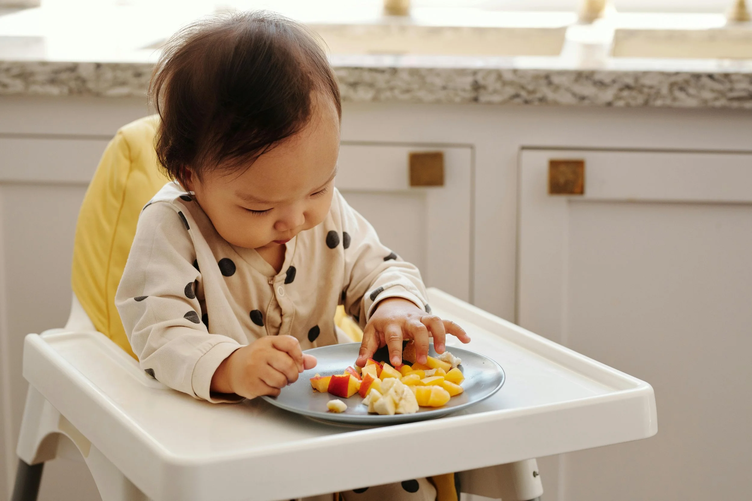 A young child with dark hair sitting in a high chair, looking at and reaching for a plate of cut fruit, including apples, bananas, and peaches, on a white tray in a kitchen with white cabinets and a granite countertop in the background.