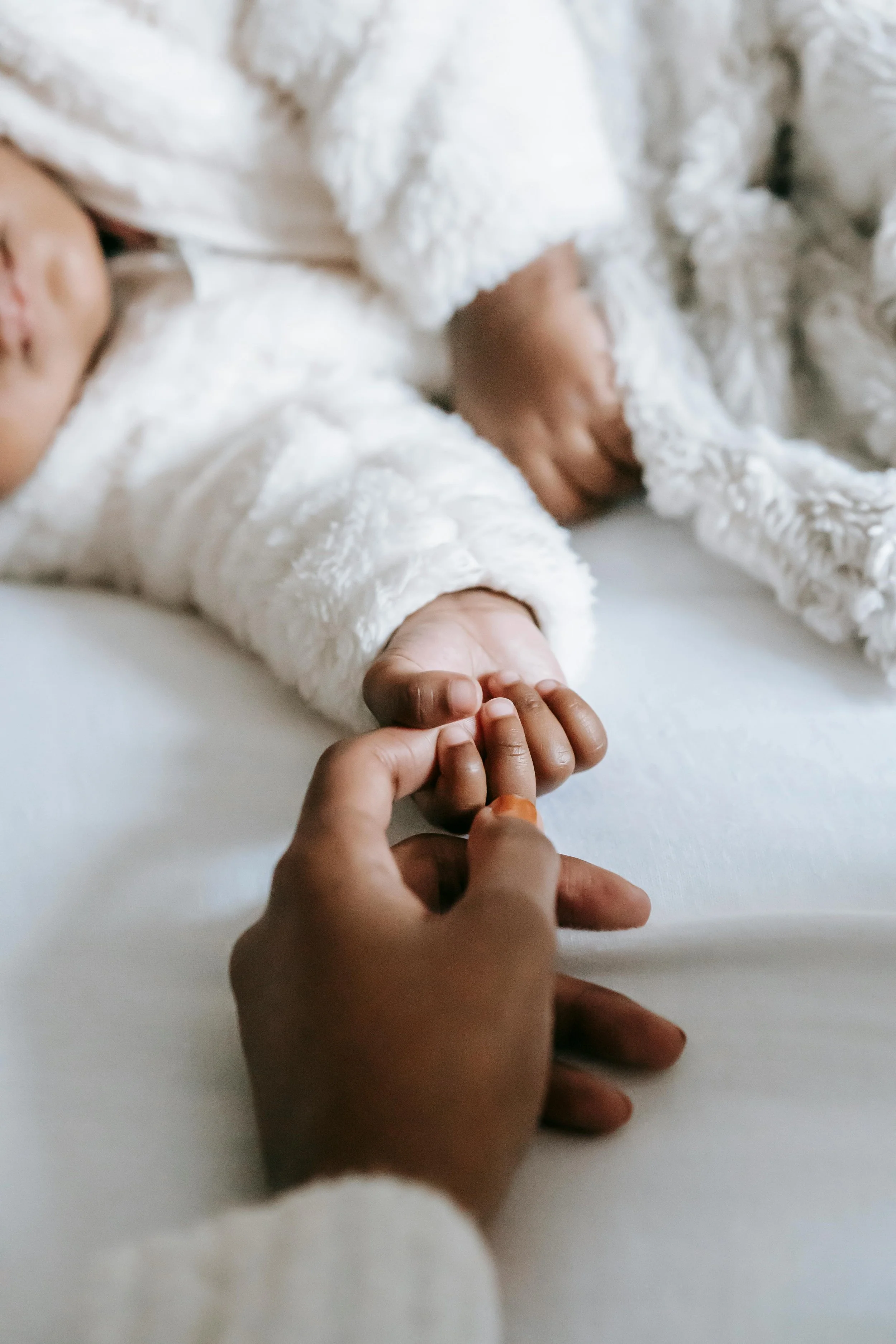 A person holding a small child's hand, both lying on a bed with white sheets. The child is wearing a fluffy white coat.