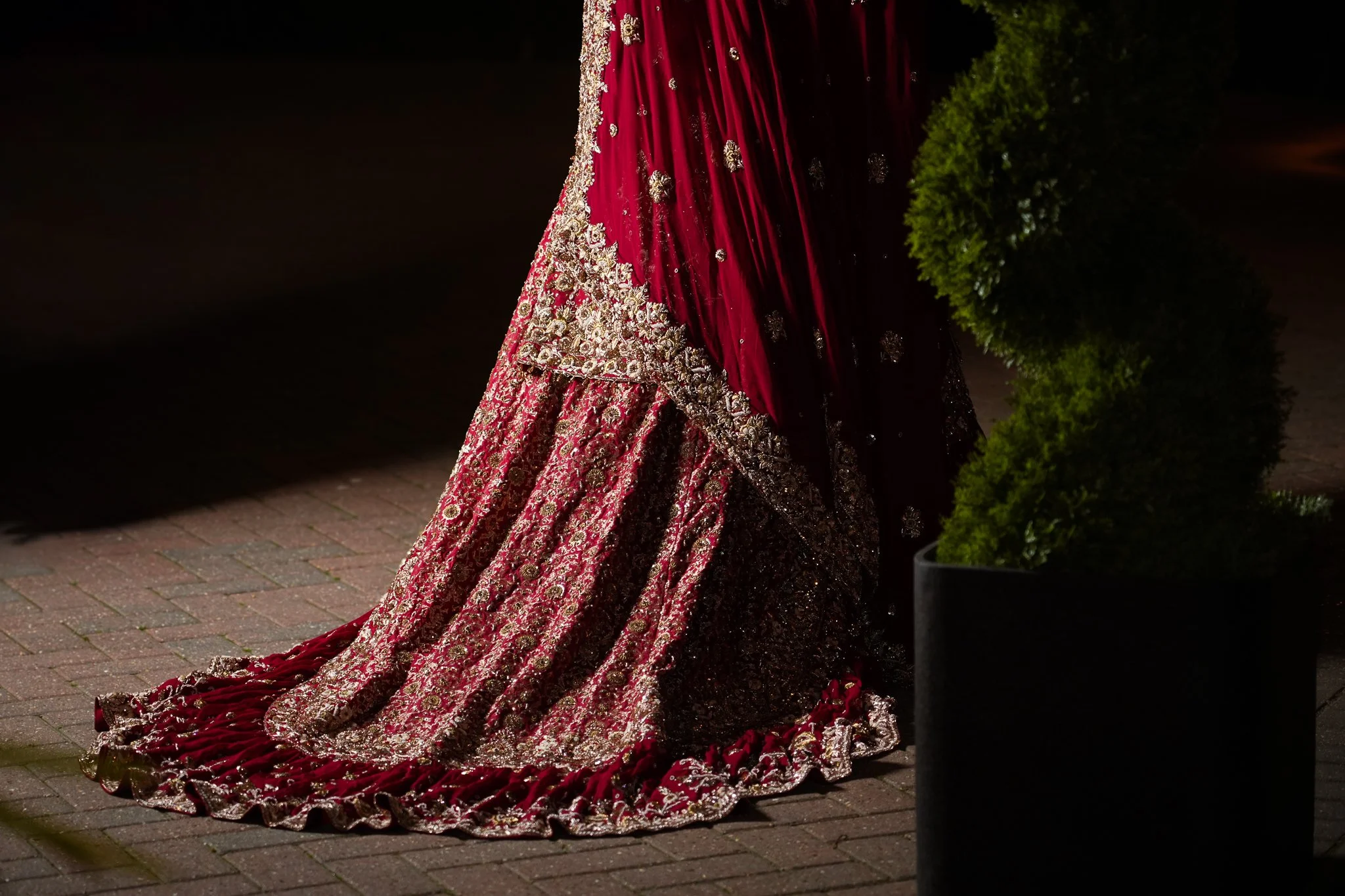 A close-up of a woman's traditional red and gold embroidered dress with a long, decorative train, standing on a brick sidewalk near a potted green shrub at night.