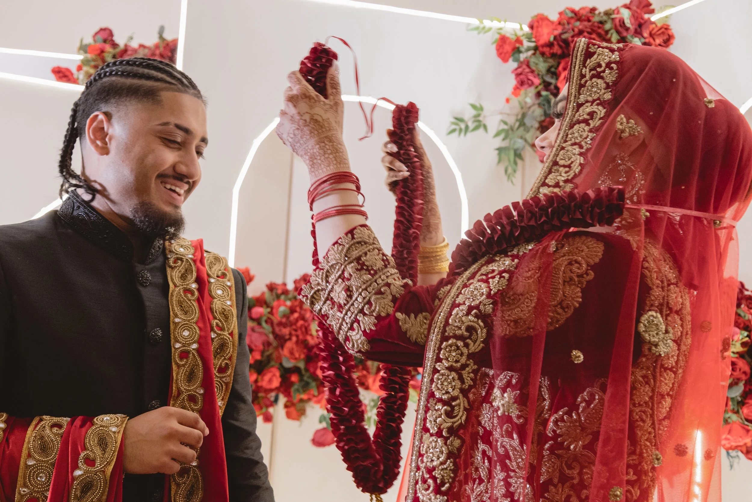 A groom in traditional Indian attire smiling as a woman, dressed in a red and gold embroidered saree with a matching red veil, holds a red floral garland.
