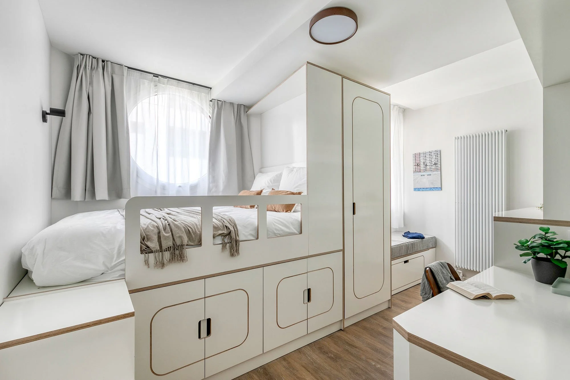 Chambre avec lit double, fenêtre ronde avec rideaux blancs, mobilier blanc avec lignes en bois, coin lecture avec lit d'appoint, décoration minimaliste avec plante verte, parquet en bois.