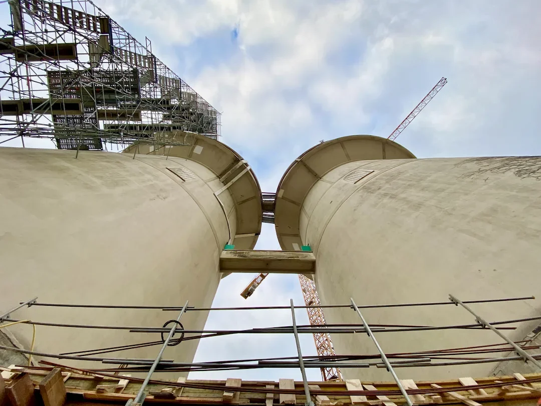 Vue à partir du bas de deux grandes silos ou tours de stockage en construction, avec une grue de chantier et un échafaudage visible, ciel partiellement nuageux.