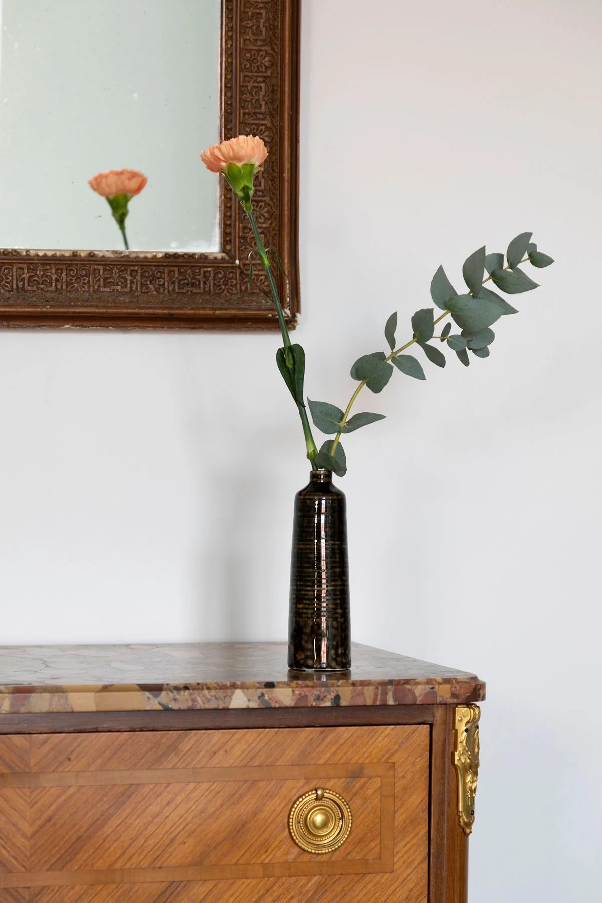 Une bouteille noire contenant deux branches de eucalyptus et une fleur de couleur pêche, placée sur une commode en bois avec une surface en marbre, au-dessus d'un mur blanc avec un miroir en bois.