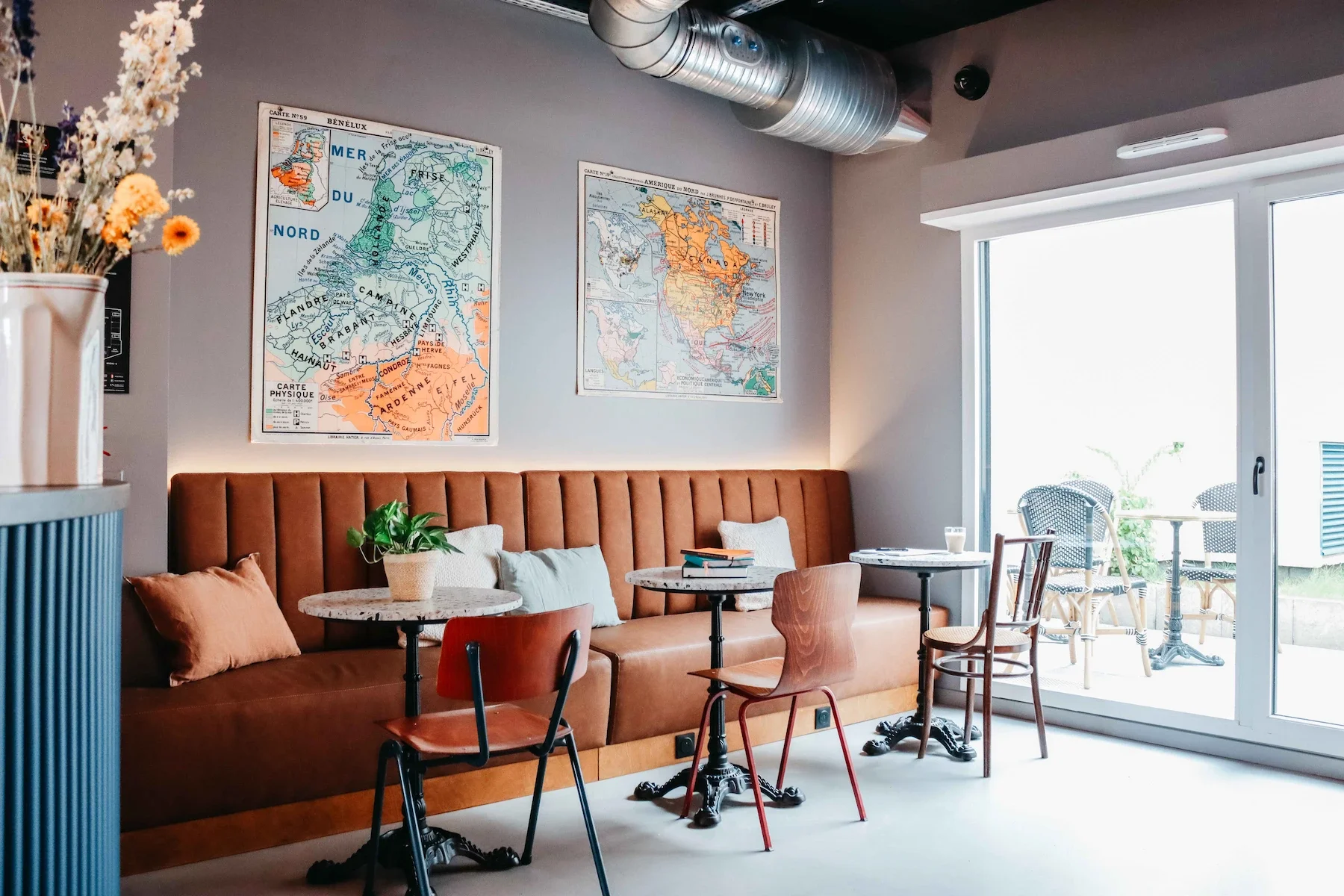 Intérieur d'un café ou d'un salon avec un canapé en cuir marron, deux cartes murales de la France et de l'Amérique du Nord, plusieurs tables avec chaises, une porte-fenêtre donnant sur un balcon avec des chaises en rotin et un peu de végétation.