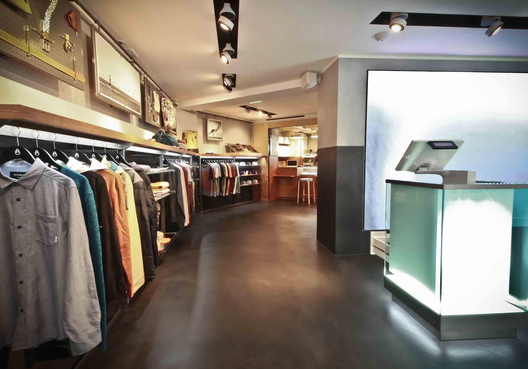 Interior d'une boutique de vêtements avec des vêtements suspendus et des articles de mode sur des étagères, une caisse enregistreuse moderne avec un écran tactile, décor contemporain, éclairage tamisé.