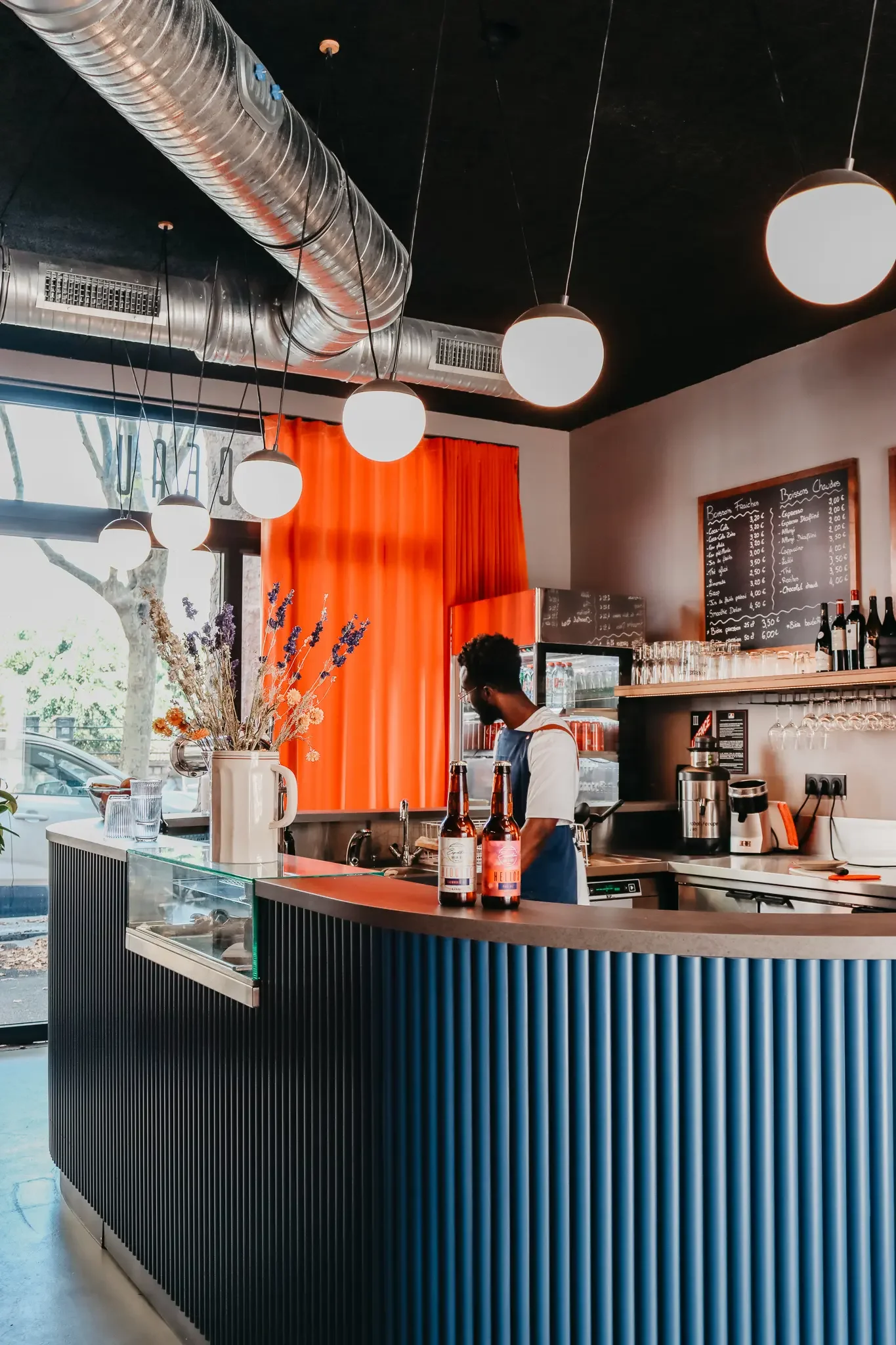Un bar moderne avec un comptoir en cuivre, une décoration florale dans une cruche, et un homme derrière le comptoir. Il y a des bouteilles de bière, une ardoise avec le menu, et des verres à vin suspendus.