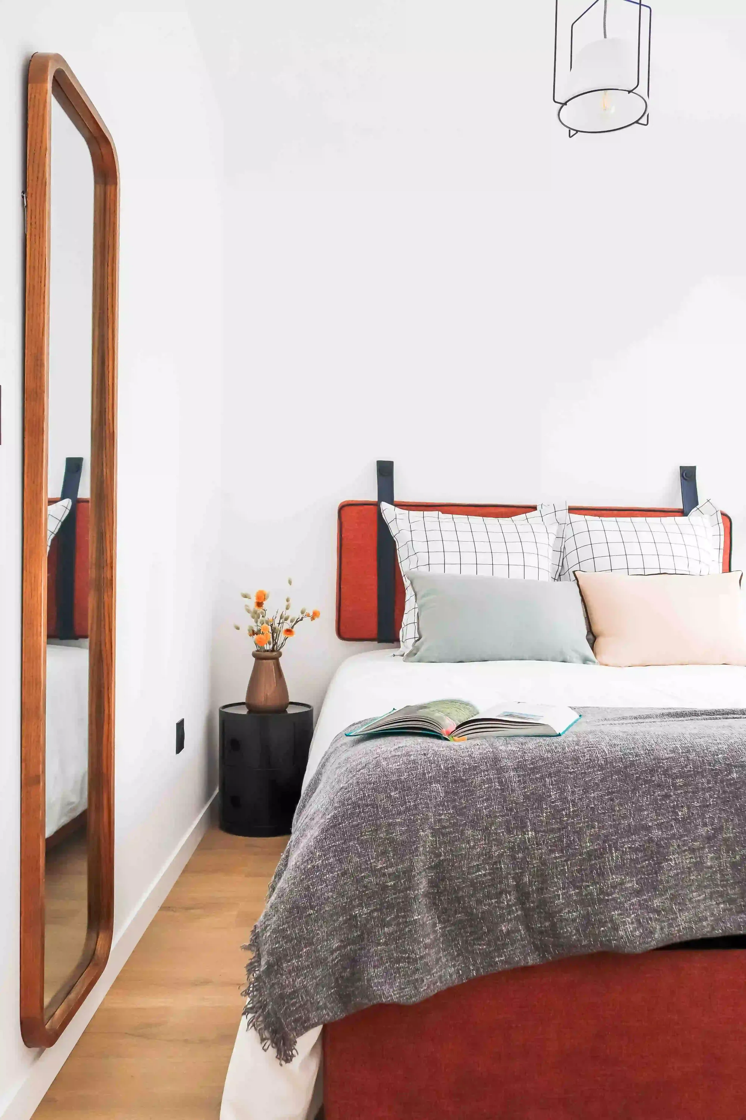 Chambre moderne avec lit à tête de lit orange, oreillers à motifs et un livre ouvert sur la couverture en tissu gris, à côté d'une petite table noire avec une vase de fleurs, miroir en bois au mur, suspension moderne au plafond.