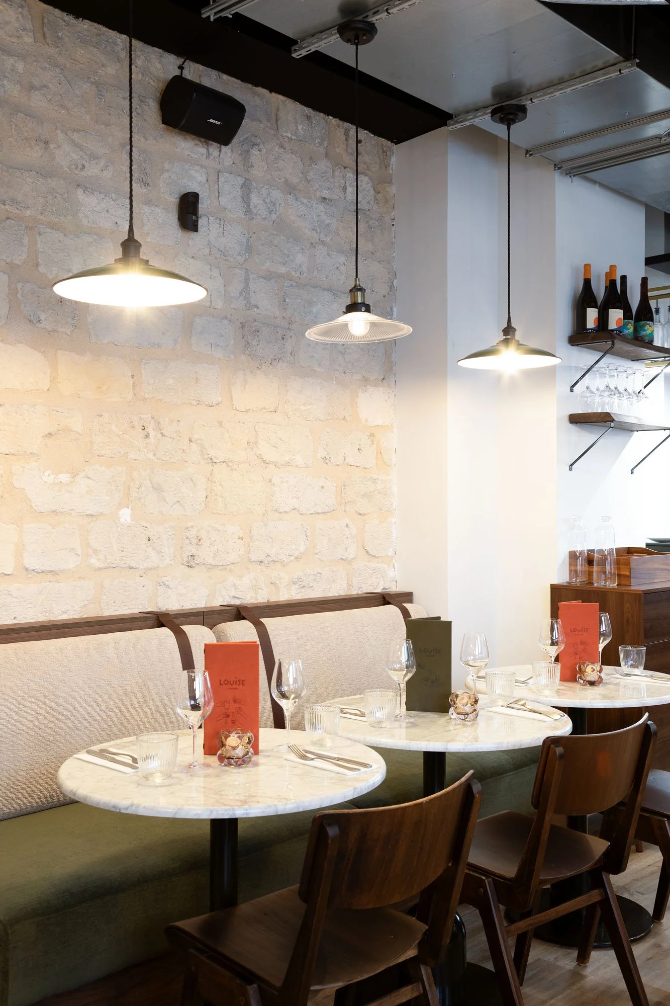 Une salle à manger dans un restaurant avec des tables en marbre blanc, des chaises en bois, des verres à vin et des menus rouges et verts sur les tables. Il y a un mur en pierre beige et des suspensions lumineuses au plafond.