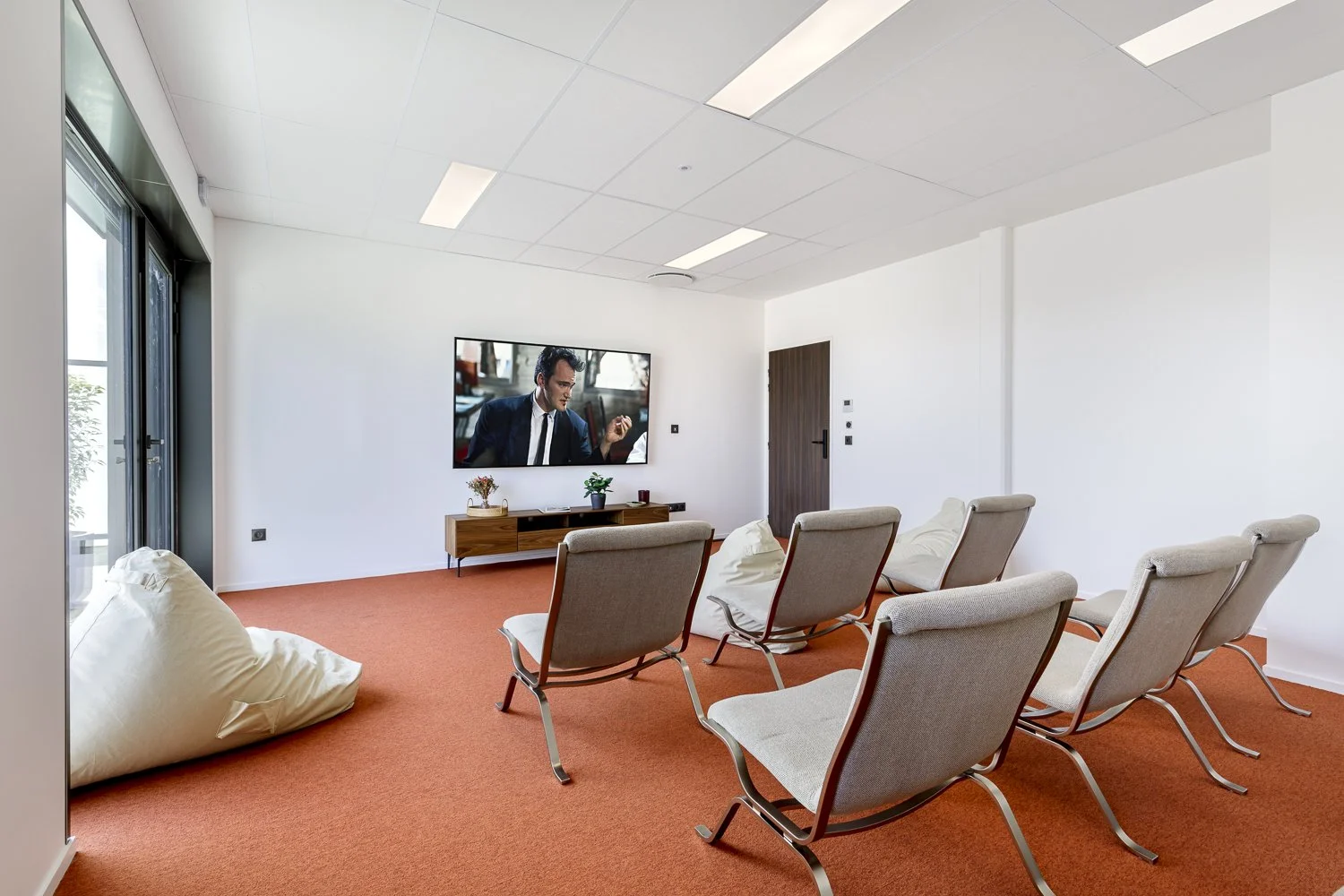 Salle d'attente avec plusieurs chaises, un grand écran TV affichant une scène avec un homme en costume, et une porte en bois. La pièce a des murs blancs, un plafond suspendu, et un sol recouvert de moquette orange.