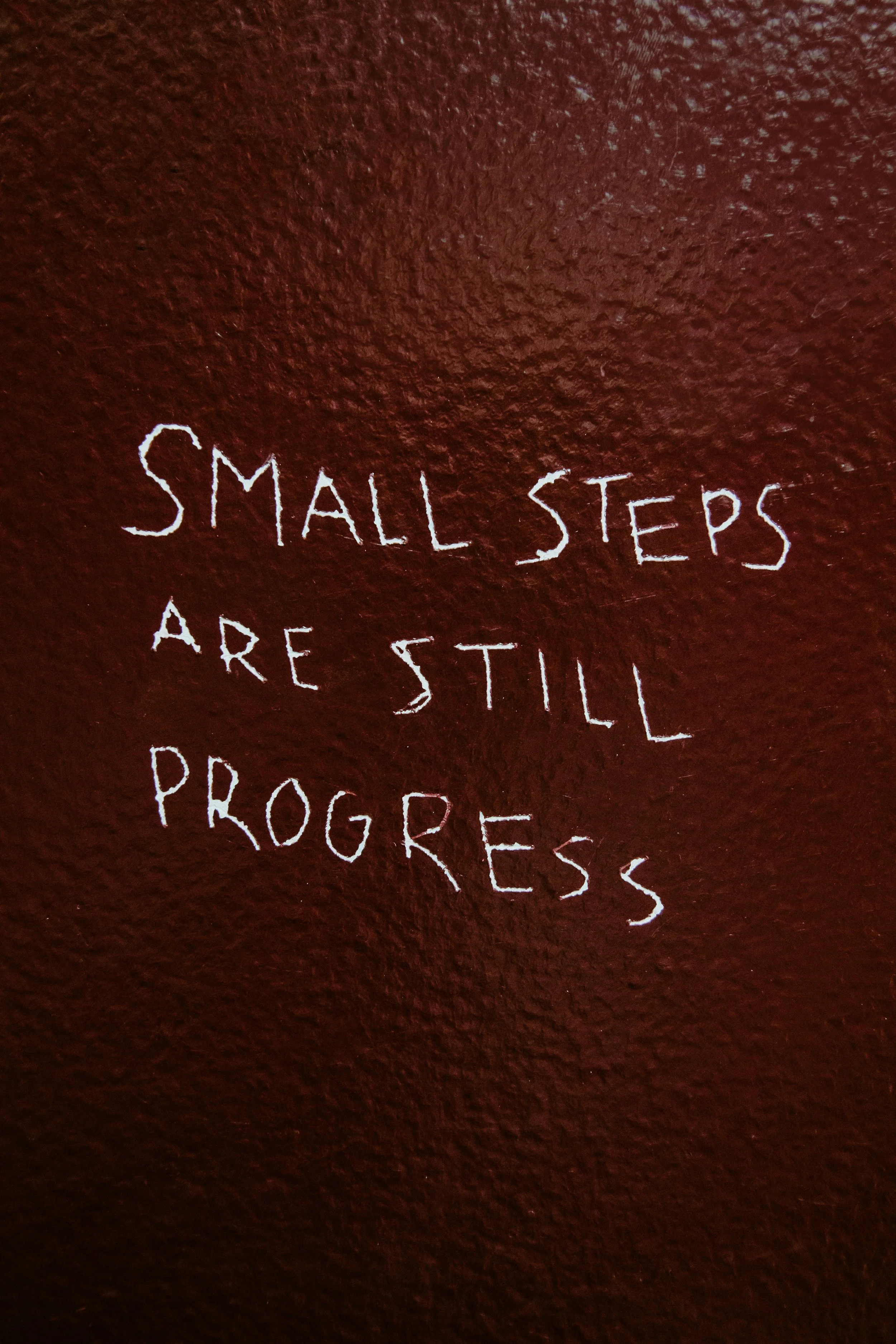 Weißer handgeschriebener Text auf einer roten Wand: 'Small steps are still progress'