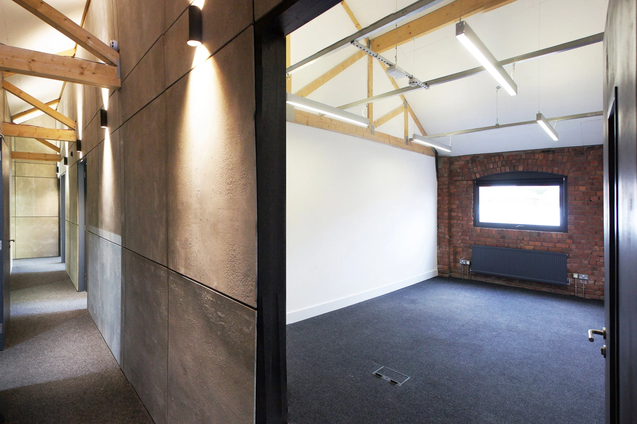 Interior of an industrial-style office with exposed brick wall, white painted ceiling with wooden beams, modern lighting fixtures, and a window with black trim.