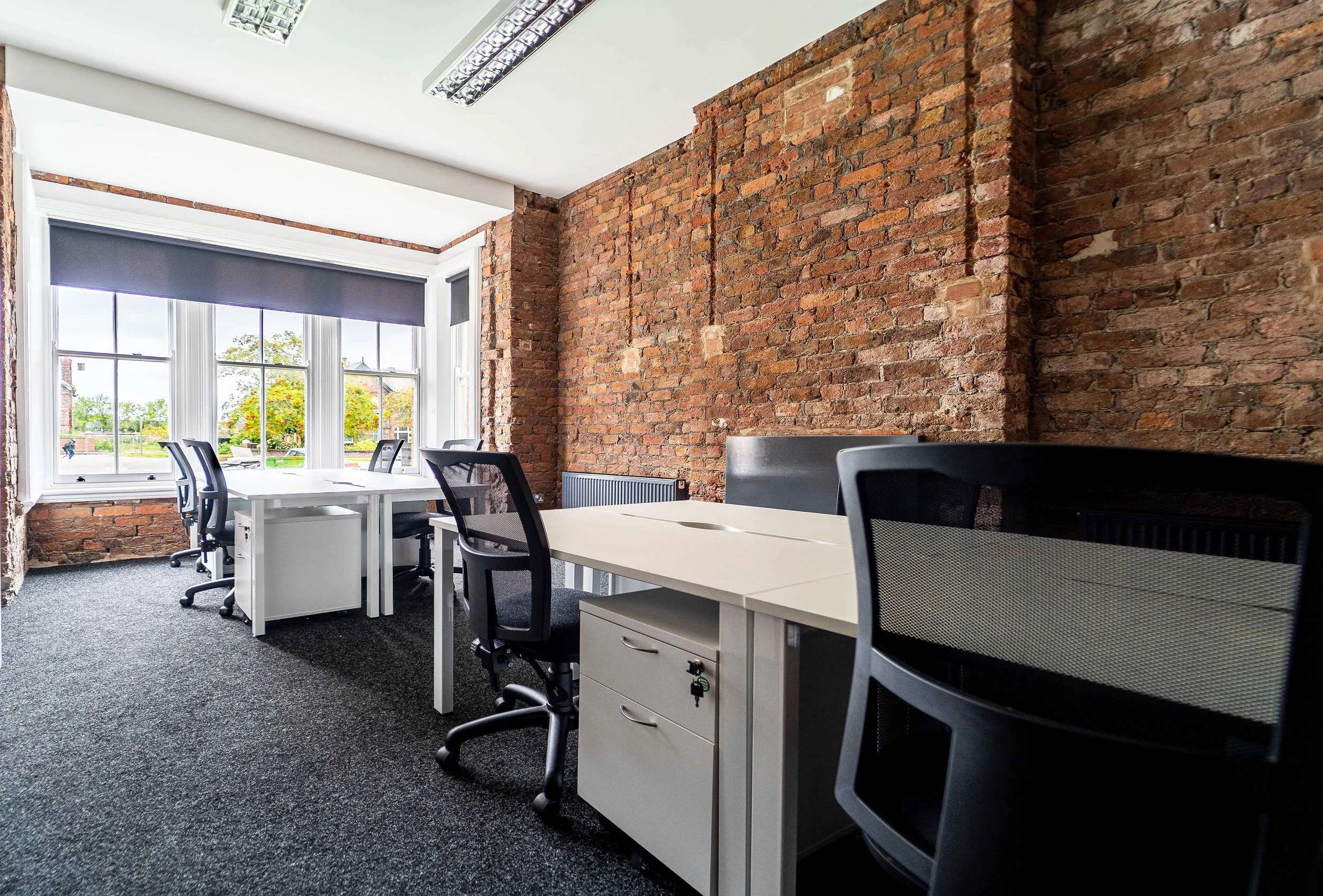 Office space with white desks and black chairs, large windows with a view of trees outside, exposed brick wall, gray carpet, and ceiling lights.